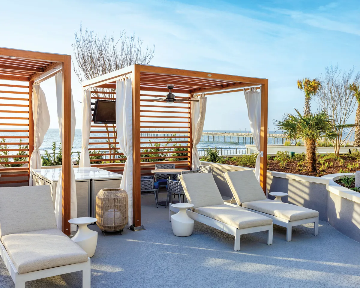 An inviting exterior scene featuring wooden cabanas and lounge chairs set along the oceanfront, with palm trees framing the view. The image captures the relaxed atmosphere of sunbathers enjoying the tranquil coastal setting, with a glimpse of the ocean in the background.