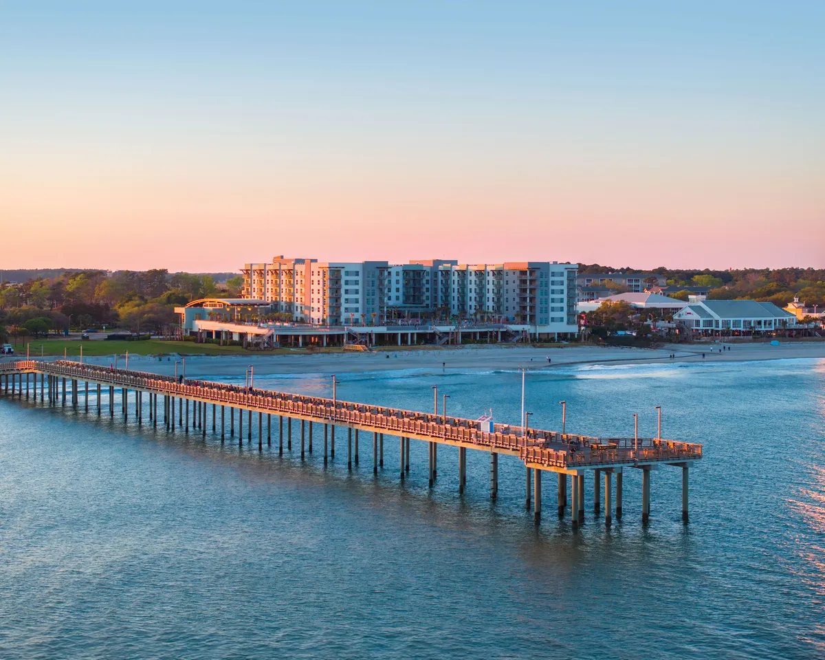 A scenic waterfront pier extends into the water beside a charming building, all framed by a vibrant sunset sky. This setting is ideal for strolls, fishing, waterfront dining, and soaking in the lively coastal atmosphere.