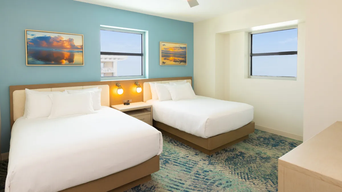 A comfortable bedroom with two queen beds, large windows, and decorative paintings. Sunlight fills the space, creating an inviting atmosphere ideal for relaxation or enjoying the view.