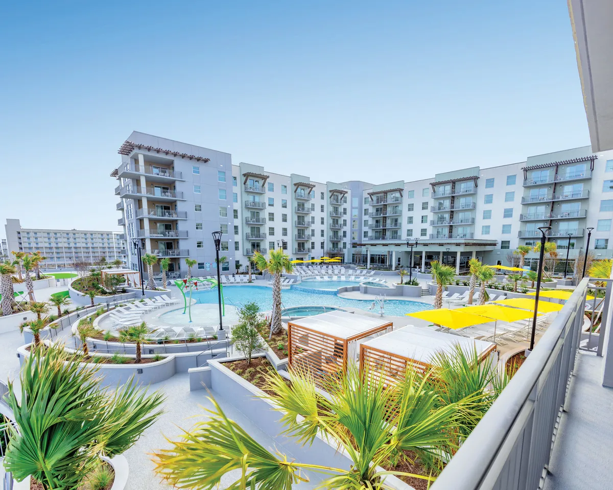 A lush pool area surrounded by swaying palm trees, vibrant yellow umbrellas, and contemporary apartment buildings. Guests lounge by the water and socialize, with the evening light casting a golden hue over the scene.