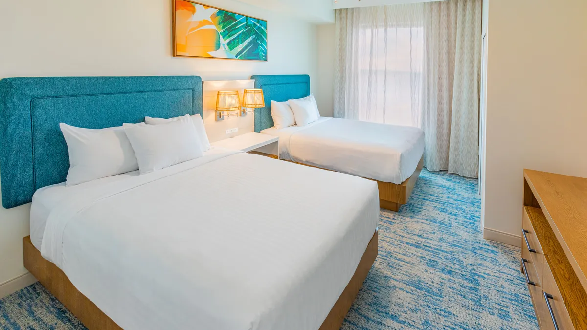 This bedroom scene features two inviting beds adorned with plush bedding, colorful artwork above the headboards, and curtains gently framing the window. Natural light streams into the room, creating a serene and welcoming atmosphere perfect for relaxation and quiet time.