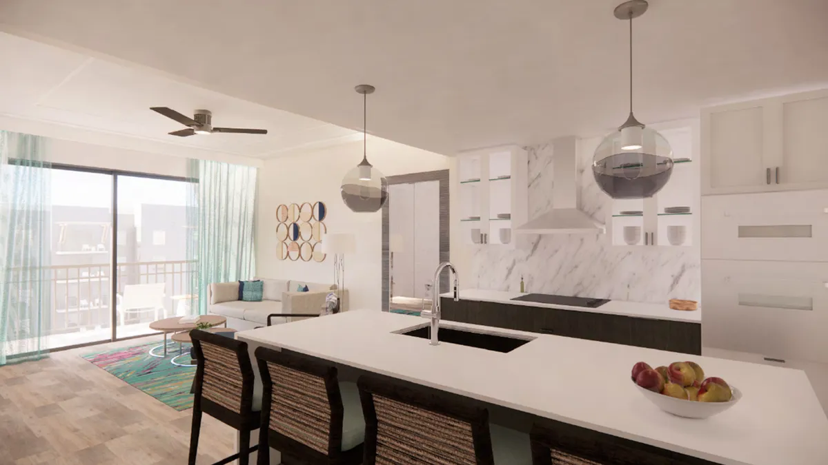 A modern, sunlit living area with an open island kitchen, decorative wall art, and a comfortable sitting area. Large windows draw in natural light and offer glimpses of the ocean beyond the balcony, highlighting the villa's elevated position.