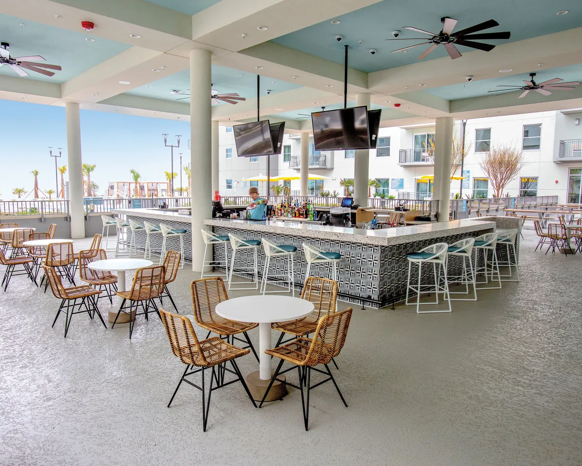 A vibrant outdoor bar area with a sleek counter, comfortable seating, and ceiling fans, all overlooking a scenic view. Guests are shown enjoying drinks and lively conversation as the sun sets, capturing the energetic yet relaxed atmosphere of the pool bar.