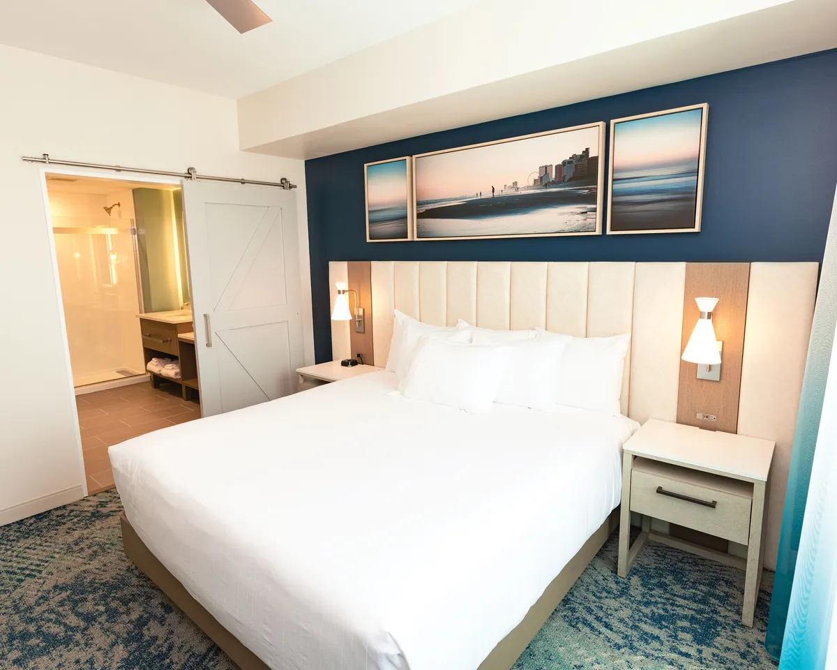 A serene bedroom featuring a plush bed, tasteful wall art, and a sliding door that likely opens to the private balcony. The tranquil ambiance is enhanced by the soft, coastal light filtering in.