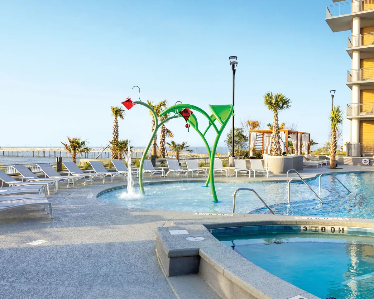A joyful outdoor pool area featuring a colorful water play structure for children, surrounded by a swimming pool, lounge chairs, and lush palm trees. Families gather to enjoy sunshine and laughter, making the space perfect for kids and adults alike.