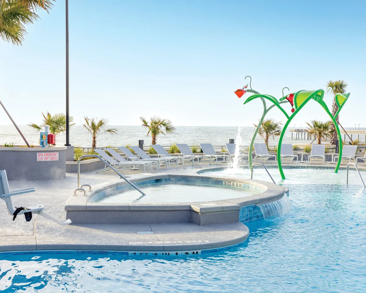 A lively oceanfront pool area features a sparkling swimming pool, an eye-catching water feature for kids to enjoy, and plenty of lounge chairs for relaxation. In the background, the ocean view sets the scene for beachfront fun and family-friendly activities.