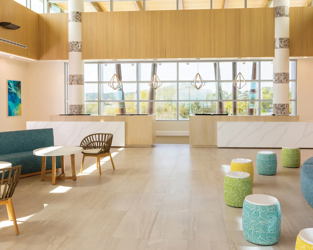 This image showcases a bright interior space with large windows flooding the room with natural light, a welcoming reception area, and colorful seating. An art piece on the wall and versatile furniture create an inviting atmosphere that encourages socializing and relaxation.