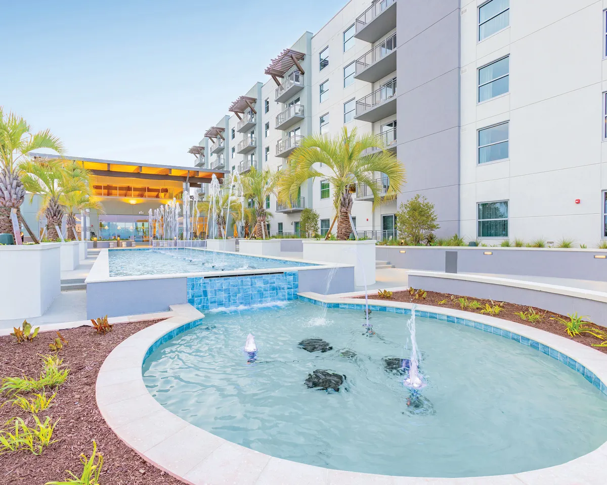 A serene exterior showcases fountains surrounded by palm trees, a modern building facade, and paved walkways inviting leisurely strolls. The area exudes a peaceful yet upscale resort atmosphere, perfect for families to gather and relax outdoors.